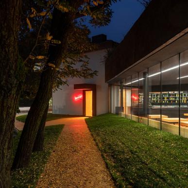 Vista notturna di un edificio vetrato con ingresso segnato da insegna al neon rossa, raggiungibile da un sentiero tra gli alberi.
