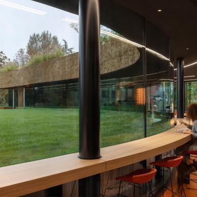 Interno con bancone curvo in legno e sedie rosse affacciato su grandi vetrate, con vista su prato e edificio rivestito di terra con tetto verde.