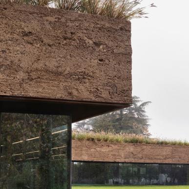 Primo piano di una parete in terra cruda con vegetazione sul tetto, sopra ampie vetrate che riflettono il paesaggio.