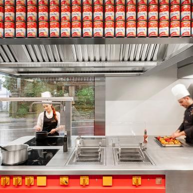 Due chef al lavoro in una cucina professionale con piano inox, pareti vetrate e scaffale decorato con file di lattine Mutti di pomodoro.
