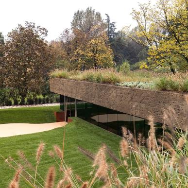 Struttura moderna parzialmente interrata, con facciata vetrata e copertura in terra e vegetazione, immersa in un parco alberato.