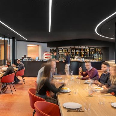 Sala ristorante luminosa con pavimento arancione, arredi in legno e sedie rosse, persone sedute ai tavoli e bancone con scaffali retroilluminati.