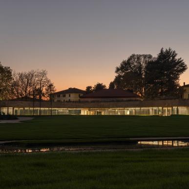 Panoramica al tramonto di un edificio con lunga facciata vetrata illuminata, circondato da prato e alberi.