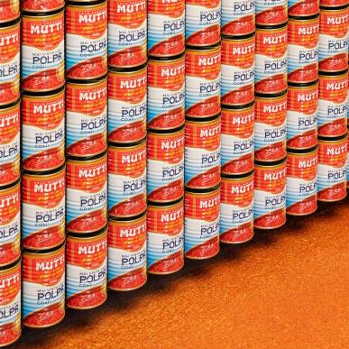 Parete rivestita da file ordinate di lattine Mutti di polpa di pomodoro, su pavimento arancione.