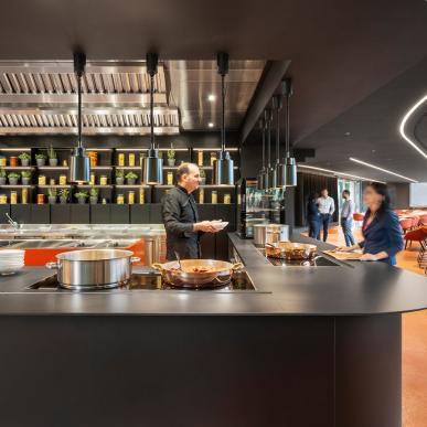 Chef al lavoro in cucina a vista con bancone nero, pentole in rame e scaffali retroilluminati con barattoli e piante.