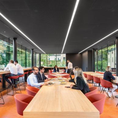 Interno con lunghi tavoli in legno e sedie rosse, persone sedute a mangiare e grandi vetrate affacciate sul verde.