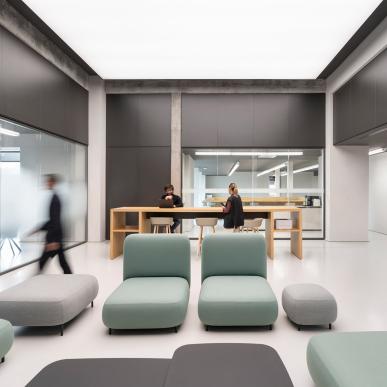 Spazio relax con divanetti moderni e area coworking con tavolo alto e persone al lavoro.