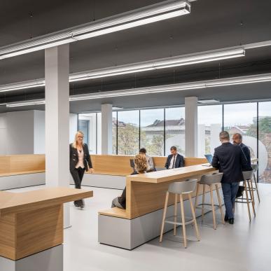 Area comune con tavoli alti e panche in legno, persone sedute e in piedi in conversazione.