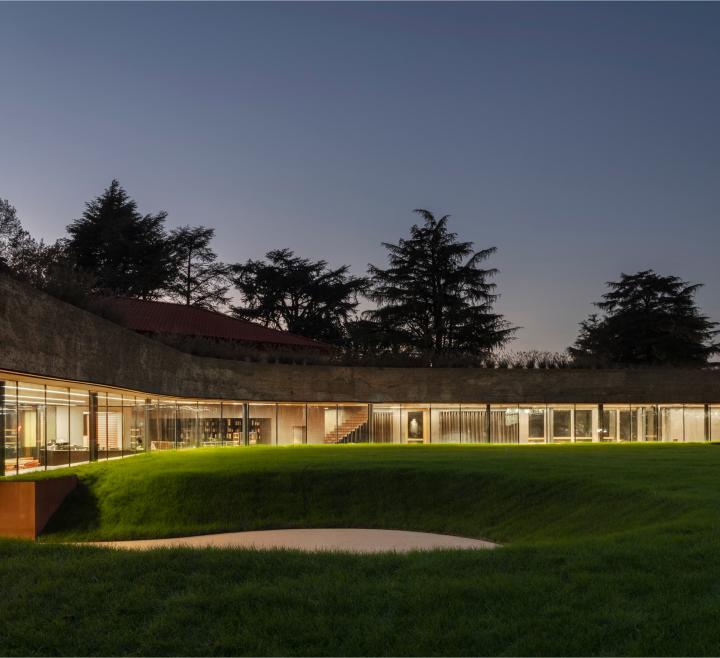 "Edificio moderno semi-interrato con ampie vetrate illuminate e prato verde in primo piano al tramonto.